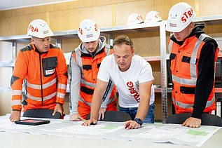 Four men in a portacabin look at a plan on the table