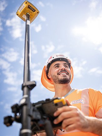 Bauarbeiter mit weissem STRABAG-Schutzhelm und Sicherheitsweste spricht per Funkgerät auf einer Baustelle. Im Hintergrund sind schwere Baumaschinen und weitere Bauarbeiter:innen im Einsatz.
