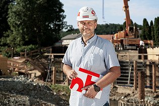 Foto Mann mit Helm und Buch steht vor einer Baugrube