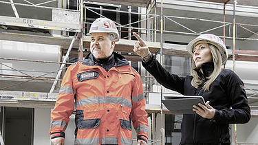 Bauführerin bei STRABAG Eine Bauführerin erklärt einem Polier etwas auf der Baustelle. Sie tragen beide STRABAG-Helme und der Mann eine orange Arbeitsjacke.
