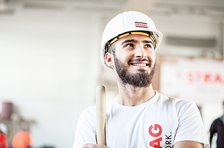 Ein lächelnder, bärtiger Bauarbeiter mit STRABAG-Helm und weissem T-Shirt hält ein Werkzeug mit Holzstiel und blickt zur Seite.