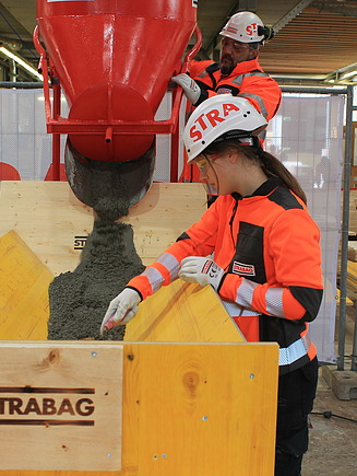 Eine STRABAG Lernende und eine Zimmermann in orangener Schutzkleidung arbeiten gemeinsam an einer Betonbefüllung. Im Vordergrund wird Beton aus einem roten Trichter in eine Holzschalung gefüllt, während die angehende Zimmerin den Beton überprüft. Im Hintergrund steht ein Zimmermann und bedient den Betontrichter. Zusätz kann man in die Werkshalle sehen, umgeben von STRABAG-Bannern und Holzverschalungen.