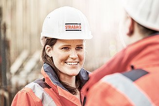 Foto Mitarbeiterin mit Bauhelm blickt Mitarbeiter mit Bauhelm an