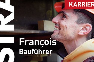 Ein STRABAG-Bauführer lächelt auf seiner Baustelle bei der Arbeit. Er trägt einen Orangenhelm und im Hintergrund kann man Stahlträger verschwommen erkennen. 