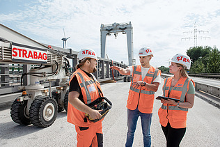 Drei Personen in Schutzausrüstung stehen auf einer Baustelle und unterhalten sich.