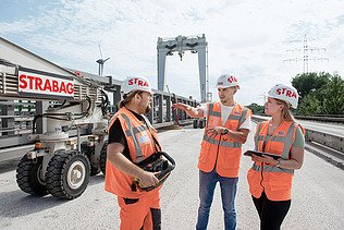Drei Personen in Schutzausrüstung stehen auf einer Baustelle und unterhalten sich.
