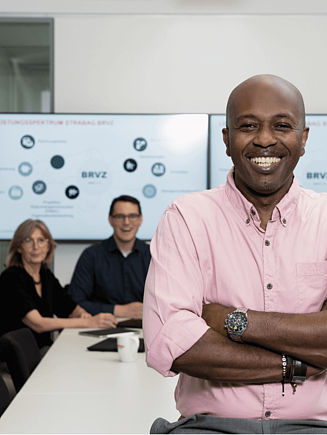 Kolega sedi na stolu u sali za sastanke i gleda u kameru, iza njega su drugi kolege