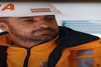 Ein STRABAG-Mitarbeiter in oranger Sicherheitskleidung mit weissen STRABAG Helm. Er ist in einem Baucontainer zeigt auf einen Plan. 
