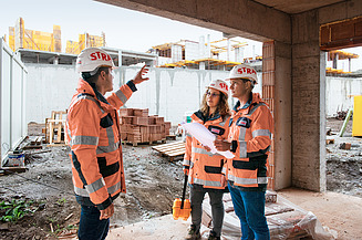 Drei Bauarbeiter:innen mit Sicherheitskleidung auf einer Baustelle, einer hält ein Dokument.