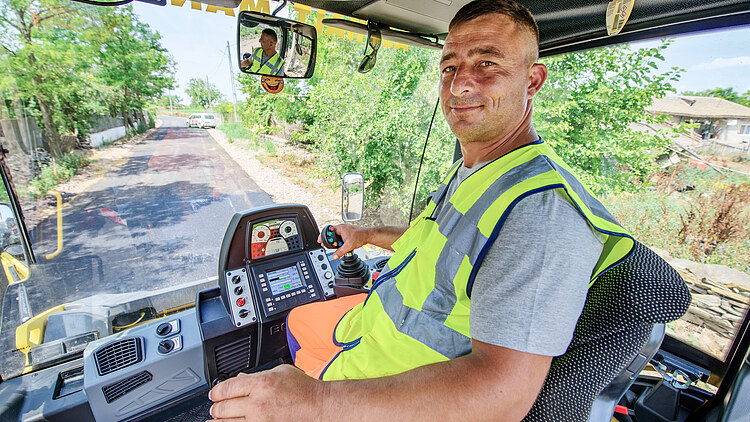 Un bărbat conduce un excavator și privește în cameră.