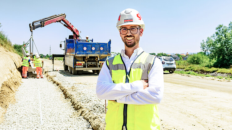 Un bărbat stă pe șantier și zâmbește către cameră.