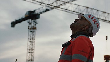 STRABAG munkatárs az építkezésen az égre néz, háttérben darukkal