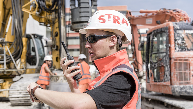 Ein Bauarbeiter mit STRABAG-Helm und Sonnenbrille spricht in ein Funkgerät auf einer Baustelle. Im Hintergrund sind mehrere Baumaschinen.