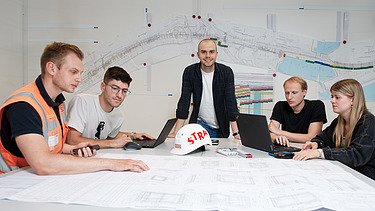 Vier Personen sitzen um einen Tisch herum, während ein Mann an der Mitte des Tisches steht und in die Kamera lächelt.