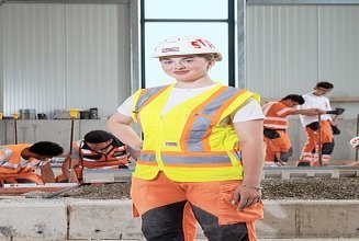 Auszubildende steht im Vordergrund mit Arbeitskleidung, während im Hintergrund weitere Lehrlinge arbeiten
