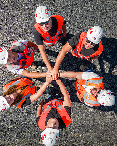 6 people in workwear and with helmets are standing in a circle, joining their right hands together in the centre. The picture is from birds-eye perspective.                                                 
