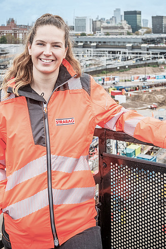 woman in workwear standing in a heightened position at a fence. A construction site is seen in the background.        