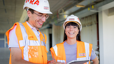 Eine Bauführerin erklärt einem Polier etwas auf der Baustelle. Sie tragen beide STRABAG-Helme und der Mann eine orange Arbeitsjacke.
