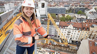 Foto von einer Bauleiterin, die auf einem Bau-Kran steht und auf die Baustelle zeigt