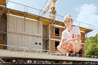 Photo: An apprentice sits in a squatting position on a structural element and looks into the camera