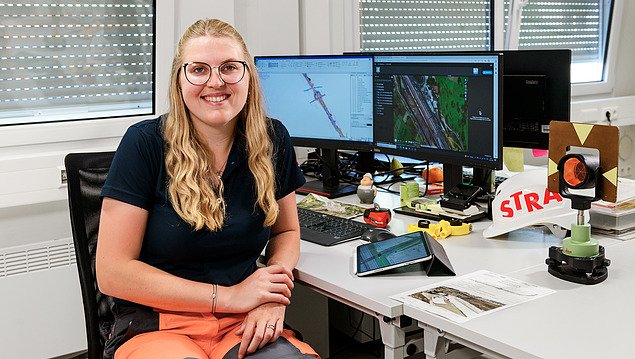 Frau in orangener Arbeitshose sitzt am Schreibtisch und lächelt in die Kamera.