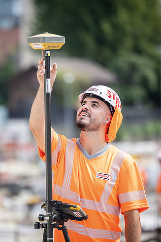 Ein lachender STRABAG Polier steht am Tachimeter auf der Baustelle.