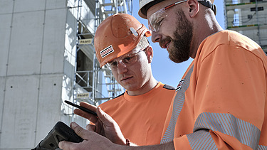 Zwei Bauarbeiter in Schutzkleidung und Helmen, einer mit STRABAG-Logo, blicken gemeinsam auf ein Tablet. Im Hintergrund sieht man einen sehr hohen Rohbau.